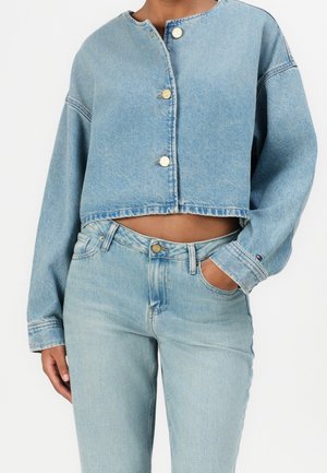 Personne portant une veste courte en denim bleu clair avec des boutons dorés et un jean taille haute assorti, une main dans la poche, sur fond blanc.
