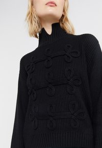 Pull en maille côtelée noire avec col montant, présentant des boucles en relief sur le devant. Texture douce et coupe décontractée.