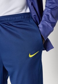 Nike trenirke u tamnoplavoj boji s teksturiranim završetkom, s elastičnim pojasom i malim žutim logom na lijevom bedru.