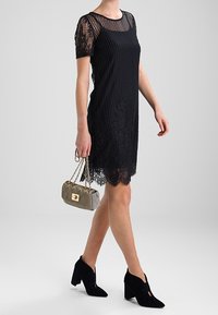 Czarna sukienka w paski z koronkowymi, krótkimi rękawami i falbaniastym dołem, zestawiona z małą metaliczną torebką i czarnymi zamszowymi botkami na wysokim obcasie.