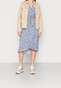 Veste en jean beige portée sur une robe bleue à fleurs avec un nœud à l'avant, présentant un ourlet à volants, associée à des baskets beige et blanches avec des accents orange.
