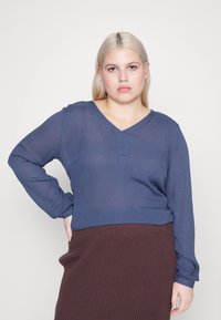 Námořnická modrá blůza s dlouhým rukávem, výstřihem do V a texturovanou látkou, v kombinaci s hnědou sukní s žebrováním. Jednoduchý design s minimem detailů.