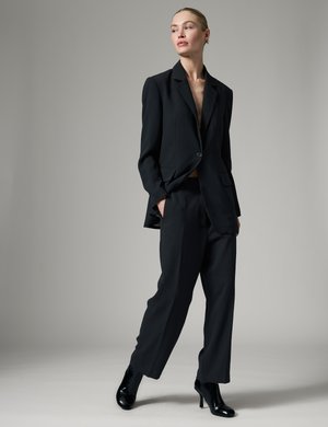 Femme debout, les mains dans les poches, portant un blazer noir oversize, un pantalon assorti et des bottines noires à talons sur fond gris.