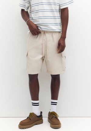 Egy férfi bézs színű cargo rövidnadrágot, fehér zoknit fekete csíkokkal, barna fűzős cipőt és fehér kék csíkos pólót visel.