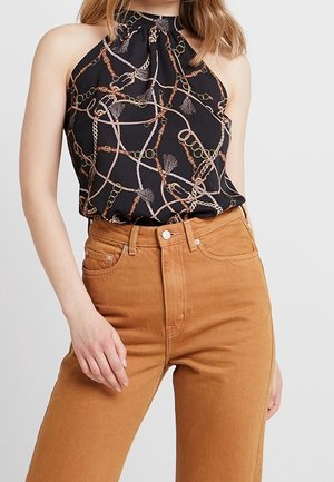 Czarny top na szyję z beżowo-brązowym nadrukiem łańcuchów i frędzli, w połączeniu z jasnobrązowymi jeansami z wysokim stanem na stojącej osobie.