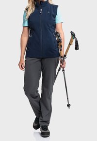 Námořnická softshell vesta přes světle modrou košili, spárovaná se šedými kalhotami. Drží trekingové hole s korkovými rukojetěmi. Černé turistické boty.