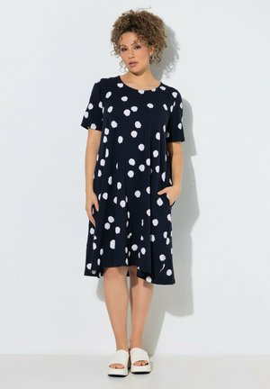 Kvinde med krøllet hår iført en sort knælængde kjole med hvide prikker, stående med den ene hånd i lommen og hvide åbne sko.