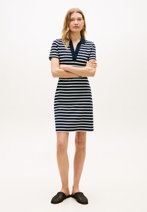 Ženska stoji z rokami prekrižanimi, oblečena v mornarsko modro in belo črtasto kratko ročno obleko in črne pletene obežne čevlje na enobarvnem ozadju.