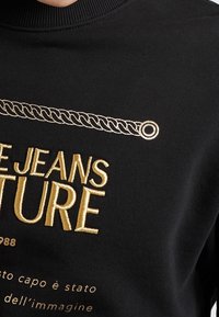 Sudadera negra con bordado dorado y diseño de cadena decorativa, con las palabras "JEANS FUTURE" destacadas.