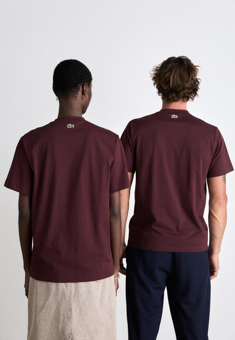 Camisetas de algodón burdeos mostradas desde la espalda, con cuello redondo y pequeños logotipos bordados en la nuca. Diseño sencillo, mangas cortas.