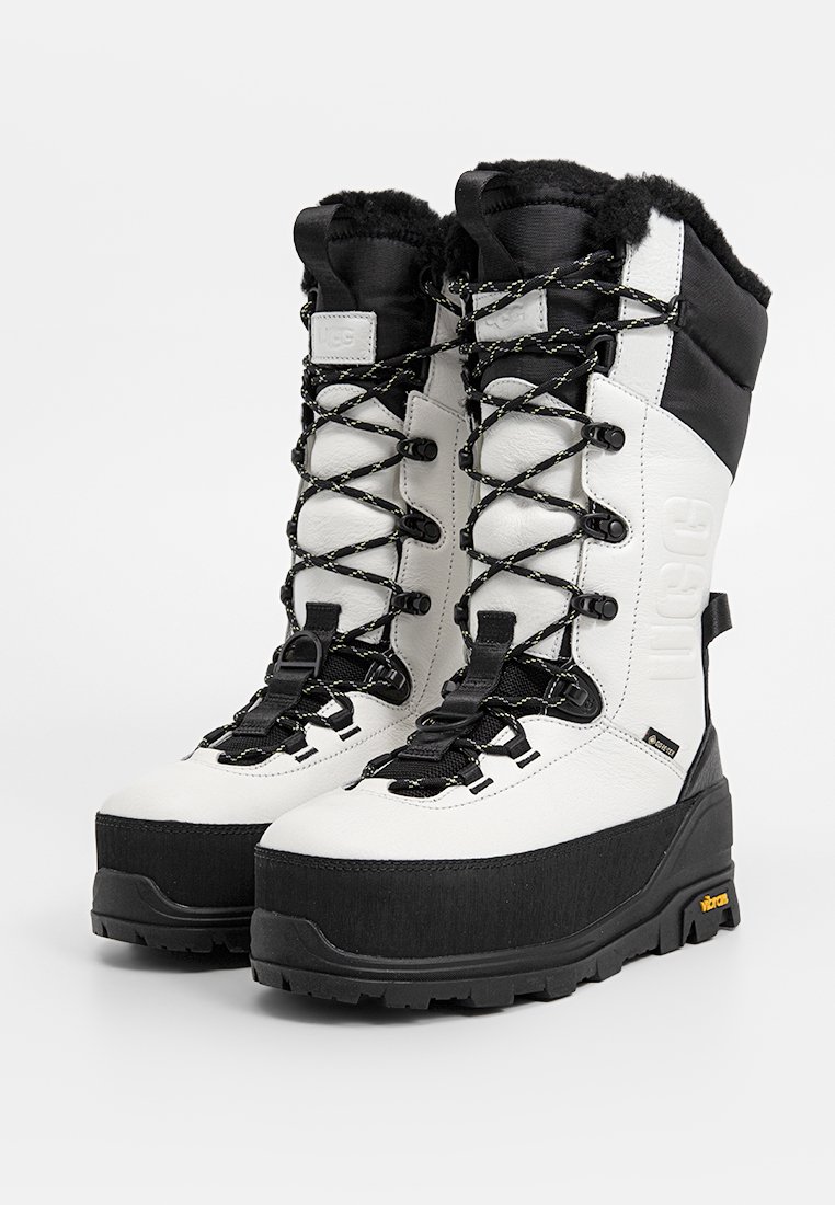 Weiße und schwarze Winterstiefel mit strukturiertem Obermaterial, dicker Sohle und runden Schnürsenkeln. Sie verfügen über ein Kunstfellfutter und ein auffälliges Logo an der Seite.