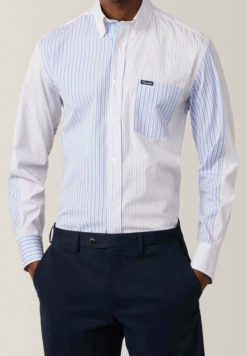 Homme portant une chemise blanche à boutons avec des rayures verticales bleues et rouges et une poche, associée à un pantalon bleu marine foncé, les mains dans les poches.