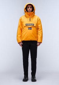 Žlutá mikina s kapucí, přední kapsou, zipovým zapínáním a logem. Nosená s černými kalhotami a černými botami.