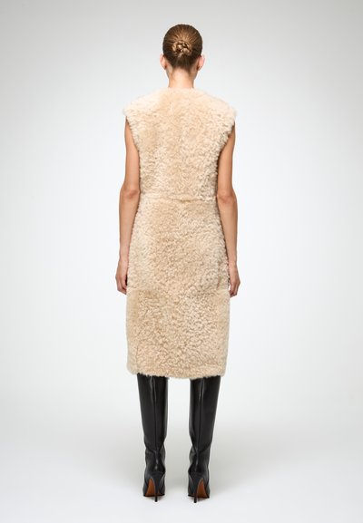 Gilet in shearling beige con una finitura morbida e texturizzata, design senza maniche e taglio di lunghezza media, abbinato a stivali neri aderenti fino al ginocchio.