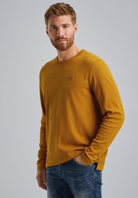 Senfgelbe Langarmbluse aus strukturiertem Material mit Rundhalsausschnitt und einem kleinen Logo-Patch auf der Brust. Getragen mit blauen Jeans.