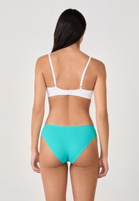 Femme aux longs cheveux bruns portant un soutien-gorge blanc et un sous-vêtement en coton ribbé turquoise, se tenant dos à la caméra devant un fond uni.
