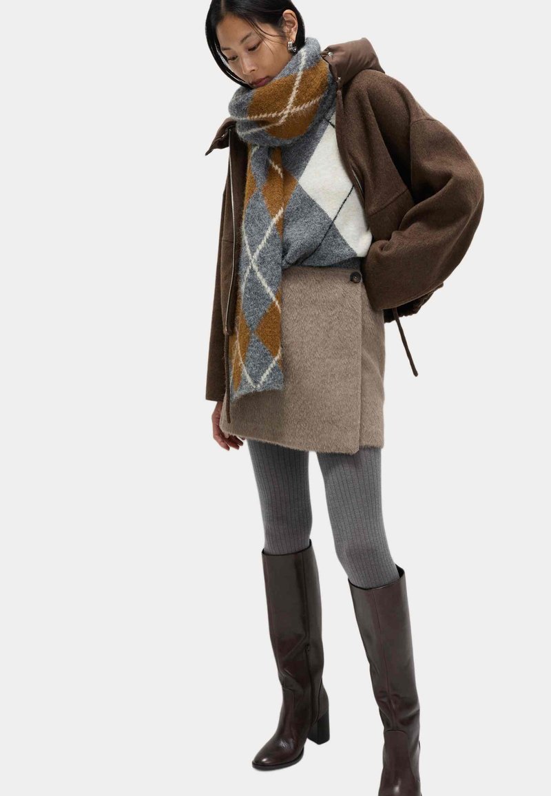 Bruine oversized jas, grijs-oranje geruite sjaal, beige getextureerde rok, grijze geribbelde panty's en bruine kniehoge laarzen.