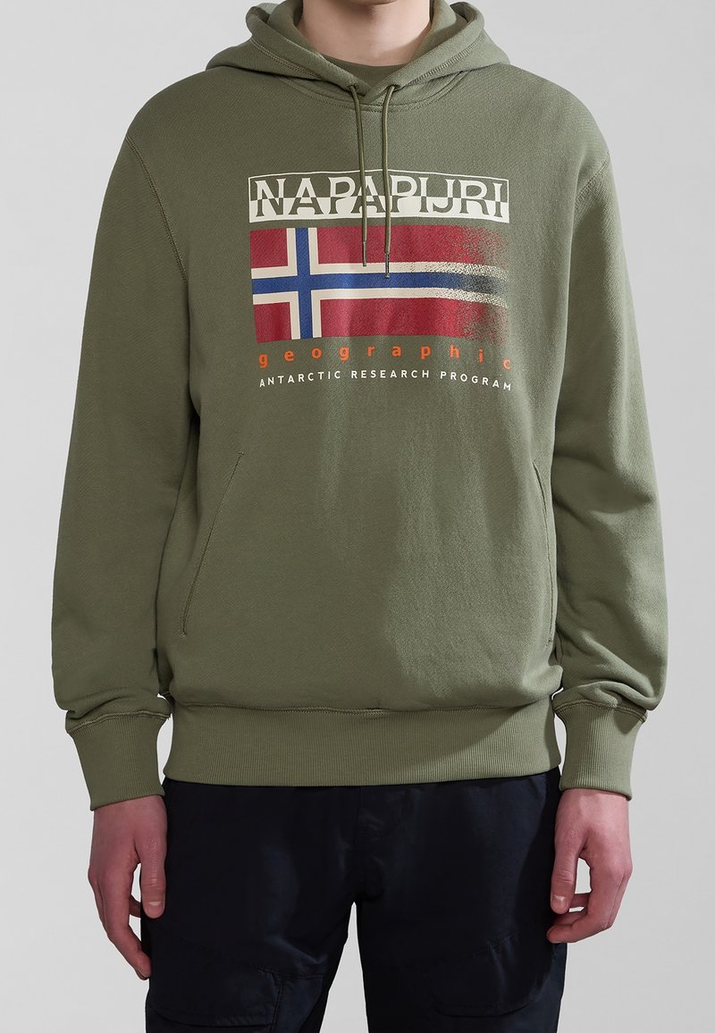 Personne portant un sweat à capuche vert olive avec poche avant, graphique du drapeau norvégien et texte "NAPAPIJRI geographic Antarctic Research Program".