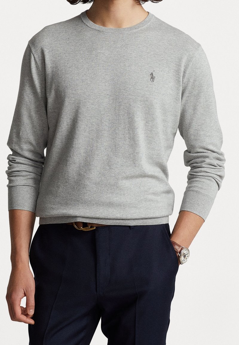 Homme portant un pull en maille gris clair à manches longues avec un petit logo brodé, pantalon bleu marine, montre à bracelet en cuir marron et ceinture marron.