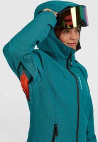 Teal technische jas met een ritssluiting aan de voorkant, verstelbare capuchon en oranje interieuraccenten. Voorzien van ritssluitingszijzakken en logodetails.