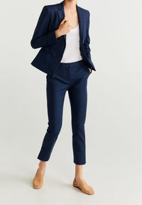 Tmavě modrý oblek s přiléhavým blejzr a na míru střiženými zkrácenými kalhotami, doplněný bílým t�ílkem a béžovými nazouvacími botami. Hladká textura látky.