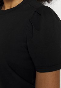 Camiseta negra de manga corta hecha de algodón. Presenta hombros abullonados y un escote de tipo crew, con costuras visibles y una textura suave.