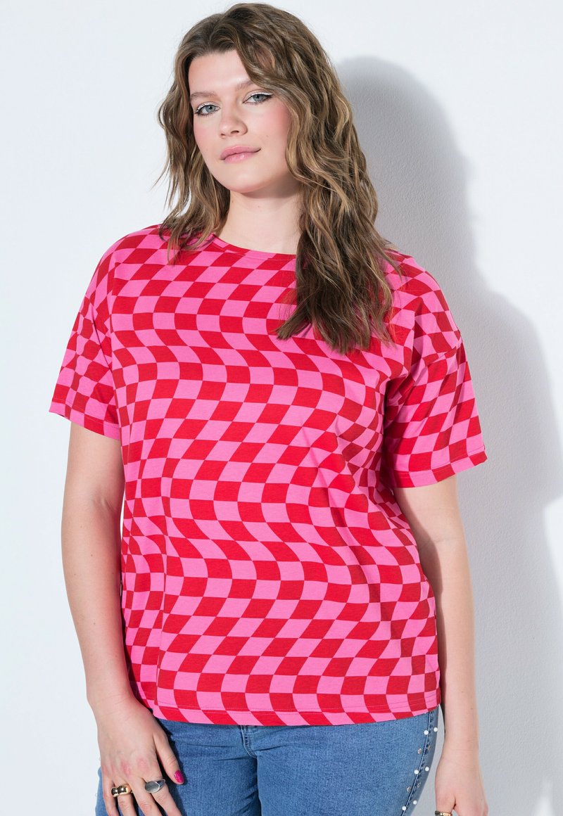 Jeune femme aux cheveux ondulés portant une chemise à motif damier rouge et rose et un jean bleu sur fond blanc.