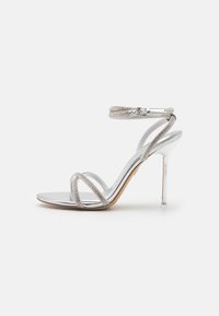 Sølv høyhælte sandal med et elegant design, med metalliske stropper og en gjennomsiktig ankelrem. Glatt metallisk finish.