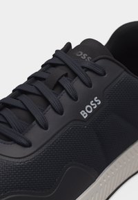 Gros plan sur une basket BOSS noire en tissu texturé, avec des lacets épais et une semelle blanche.