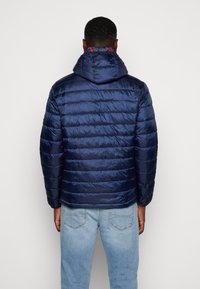 Giacca a vento blu navy con un esterno lucido e trapuntato e cappuccio, caratterizzata da un rivestimento rosso a contrasto e polsini elasticizzati per una vestibilità aderente.