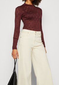 Burgundy långärmad spetsblus med rund halsringning, tillsammans med krämfärgade vida byxor, hållna av en hand som greppar en svart väska.