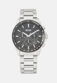 Montre en acier inoxydable BOSS avec un cadran noir texturé, des aiguilles argentées, trois sous-cadrans, une lunette tachymétrique et un bracelet en maillons métalliques.