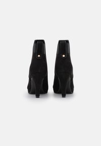 Bottines noires en suède avec des accents noirs brillants et un détail de bouton doré. Elles sont dotées d'un talon épais et haut et d'une texture lisse.