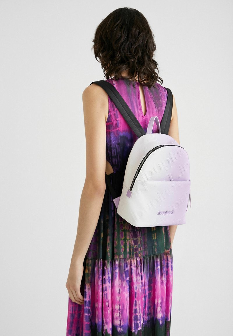 Femme portant une robe sans manches à motifs violet-rose avec un petit sac à dos blanc avec des bretelles noires et un texte en relief.