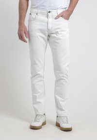 Jean en denim blanc avec une coupe slim et des coutures contrastées. Doté de poches avant et d'un zip, porté avec des baskets blanches.
