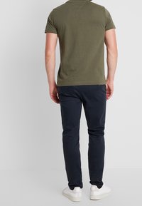 Mand i olivengrøn kortærmet t-shirt, marineblå slim-fit bukser og hvide sneakers står med ryggen til på ensfarvet baggrund.