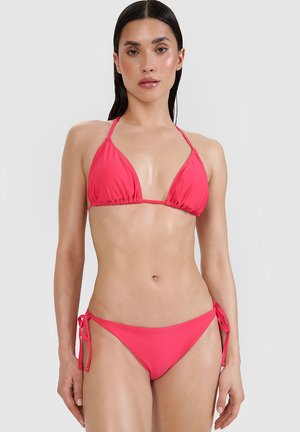 Kobieta o długich ciemnych włosach, ubrana w jaskraworóżowy top bikini halter i dopasowane bikini z wiązaniem po bokach, na jednolitym tle.