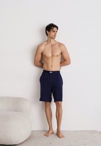 Model îmbrăcată cu pantaloni scurți bleumarin dintr-un material ușor, stând descult în fața unui perete simplu, cu mâinile împreunate în spate.