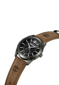 Orologio tondo nero con lancette e indicatori in tonalità argento, un quadrante secondario più piccolo e un cinturino in pelle marrone goffrato con un logo. Cassa in metallo.