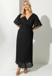 Blondynka ubrana w czarną plisowaną sukienkę midi z krótkimi falbaniastymi rękawami i metallicznym wzorem w kropki, stojąca przy białej ścianie.