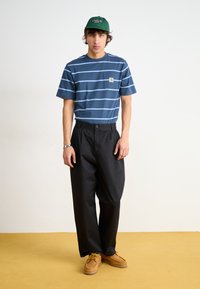 T-shirt bleu à rayures avec une poche poitrine, pantalon large noir et chaussures à lacets marron avec des accents jaunes, sur fond jaune.