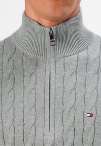 Mann mit einem grauen Strickpullover aus Kabelstrick mit hohem Rippkragen und einem halben Reißverschluss, der ein kleines rechteckiges Flaggenlogo auf der Brust trägt.