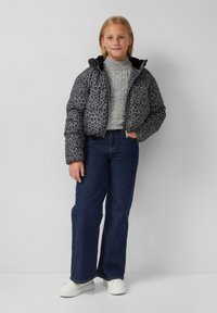 Grijze pufferjack met luipaardprint en een zwarte capuchon, gedragen over een lichtgrijze gebreide trui en blauwe flared jeans; witte sneakers.