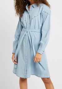 Light blue denim shirt dress with long sleeves, front buttons, decorative studs, and a tied waist for a fitted silhouette.