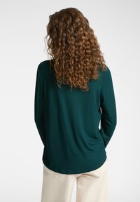 Pitkähihaista yläosaa tummanvihreästä kankaasta, jossa on sileä pinta, pyöreä pääntie ja hieman kaareva helma.