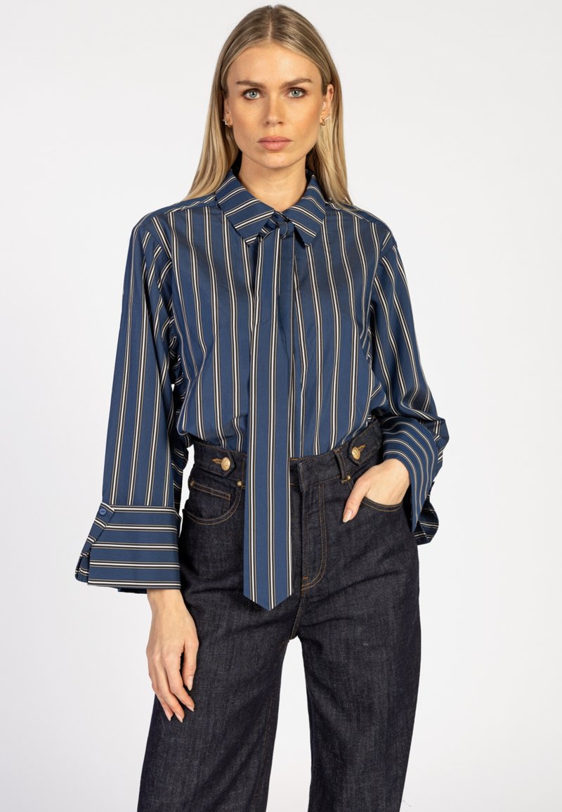 Navy striped blouse with a tie neck, long flared sleeves, and contrasting light stripes. Paired with high-waisted dark denim jeans.