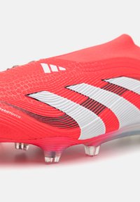 Red soccer cleat with textured synthetic upper, featuring three white stripe accents and translucent sole with visible studs.