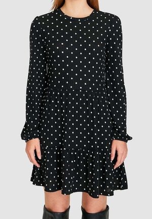 Femme portant une robe noire à manches longues avec des pois blancs, une jupe à volants et des bottes noires montantes sur un fond uni.