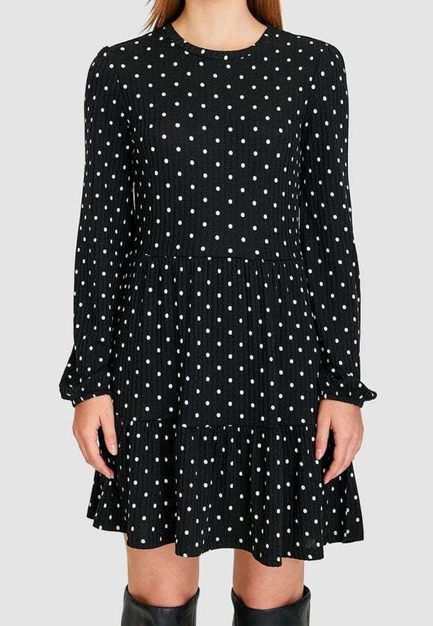 Femme portant une robe noire à manches longues avec des pois blancs, une jupe à volants et des bottes noires montantes sur un fond uni.