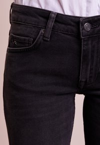 Černé džíny vyrobené z denimu s úzkým střihem, přední kapsou se stehujícími detaily a kovovým zapínáním v pase.